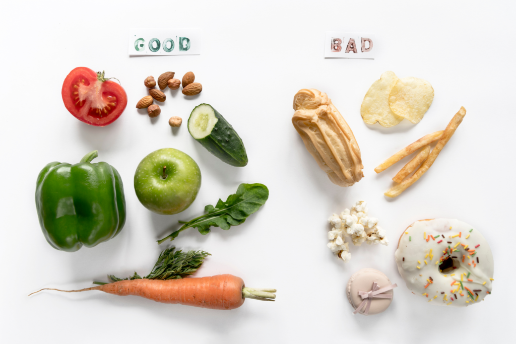 Foods lain out on a white background. on the Left side, the word "good" is written above veggies, fruit, and almonds. On the right, the word "bad" is written above chops, doughnut, popcorn, fries, and backed goods.