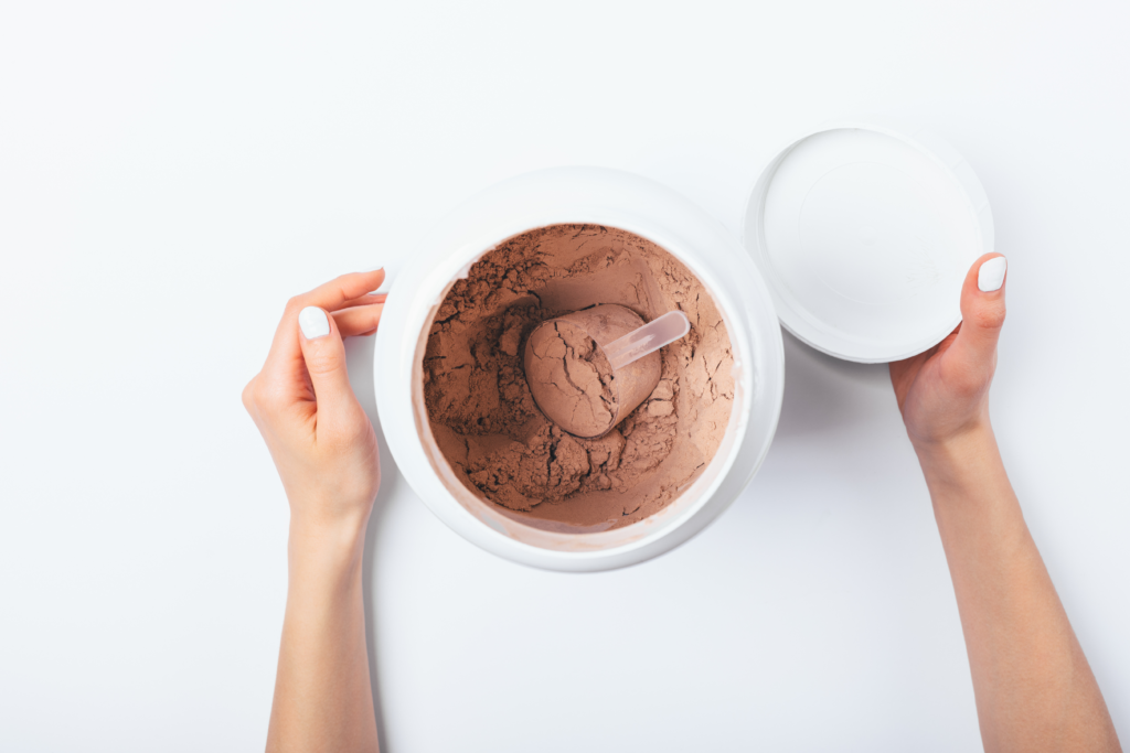 Overhead image of a container of chocolate protein powder with scoop. Woman's hands are seen opening the container