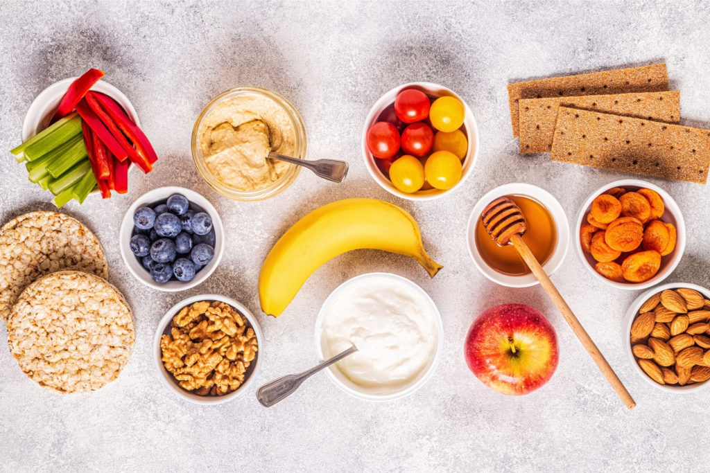 Image of overhead view of a white countertop covered in various healthy snack options like fruit, nuts, crackers, hummus and veggies