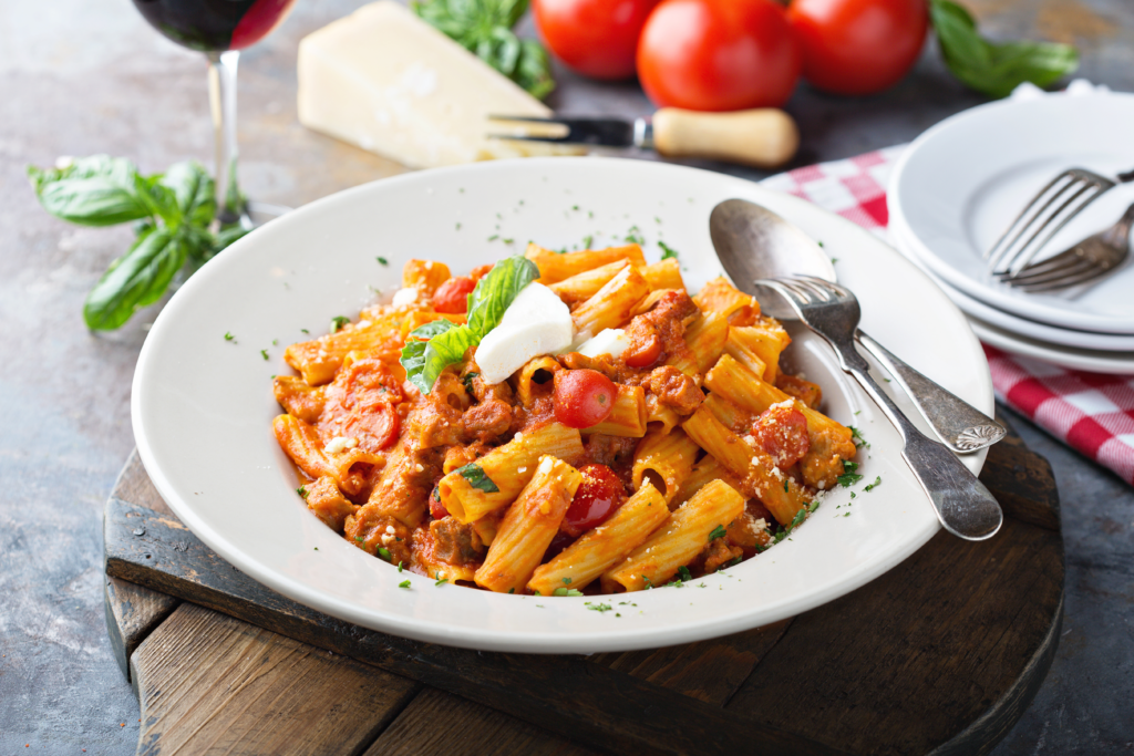 Image of rotini pasta with tomato sauce in a white bowl. Behind the bowl is red wine, tomatoes, herbs, and flatwear.