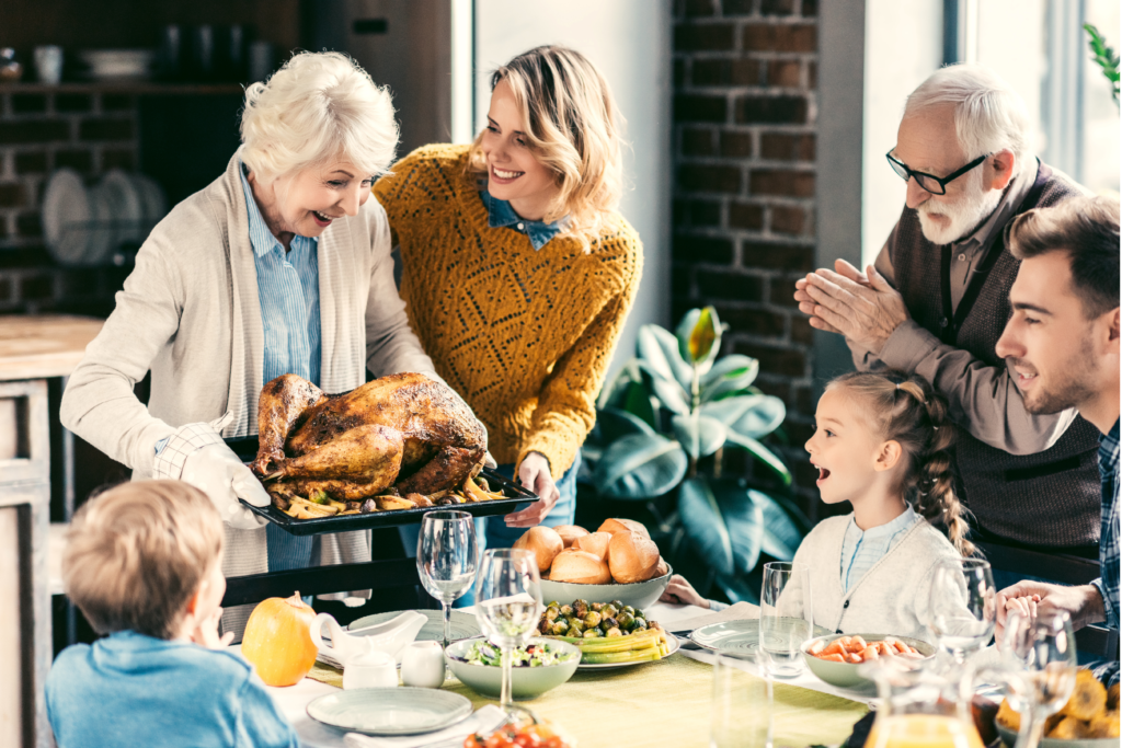 Image of family serving thanksgiving dinner in the dining room. Grandma carrying the turkey over to the table where other family members look excitedly at the turkey.