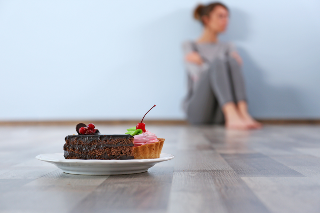 Image of food guilt. Woman sitting on floor with arms crossed. Plate of cakes on the floor in front of her that she is trying to ignore.