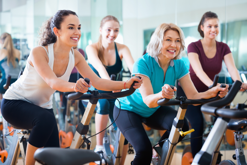 Image of four women on stationary bikes taking a cycling class. The women are smiling as they work out 