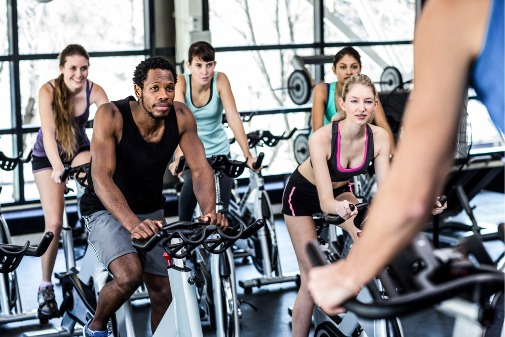 Four women and one man sitting on spin bikes facing a spin instructor as they take a workout class.