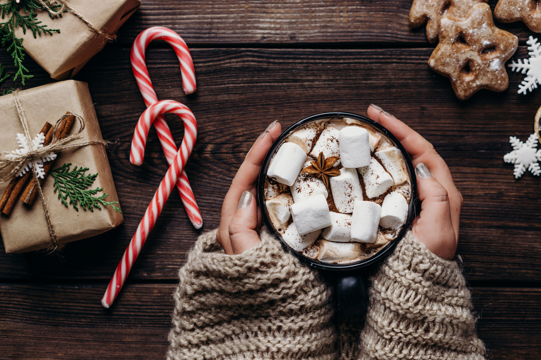 hands holding holiday hot chocolate with marshmallows on a wooden table. Candy canes and presents surround the hot chocolate on the table