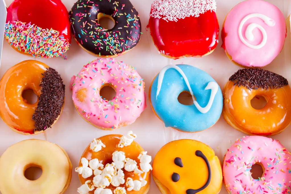 Overhead food image of a variety of doughnuts. Some are decorated with sprinkles, some with popcorn, and one with a smiley face.