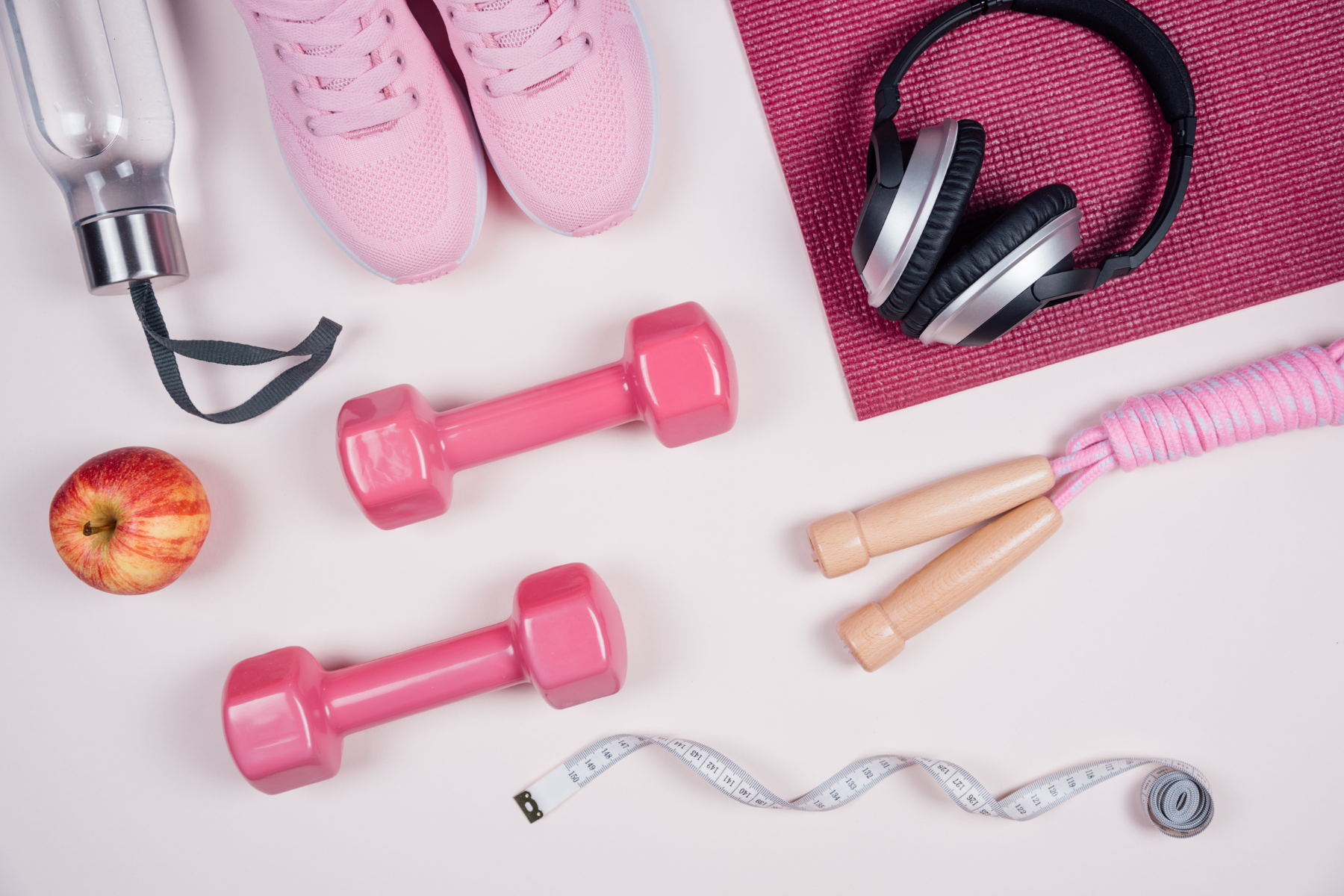 Overhead view of a pile of fitness equipment including weights, jump rope, headphones, yoga mat, running shoes, water bottle, and an apple