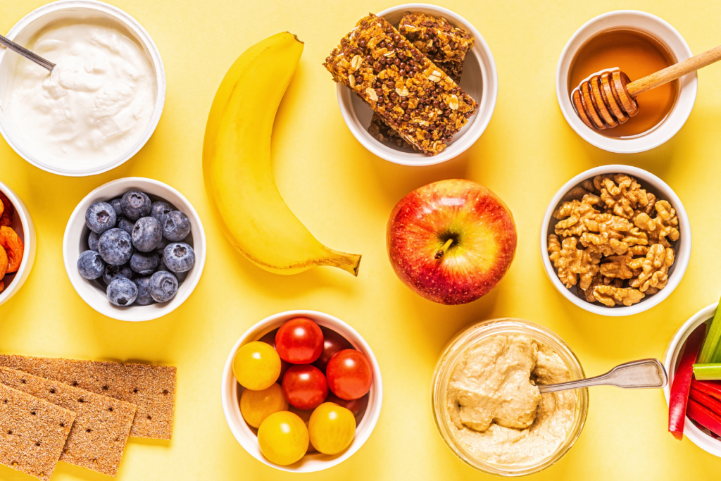 Healthy snack ideas for an active lifestyle or pre/post workout on a yellow backdrop. Apple, banana, granola bar, yogurt, hummus, walnuts, crackers, tomatoes, honey.