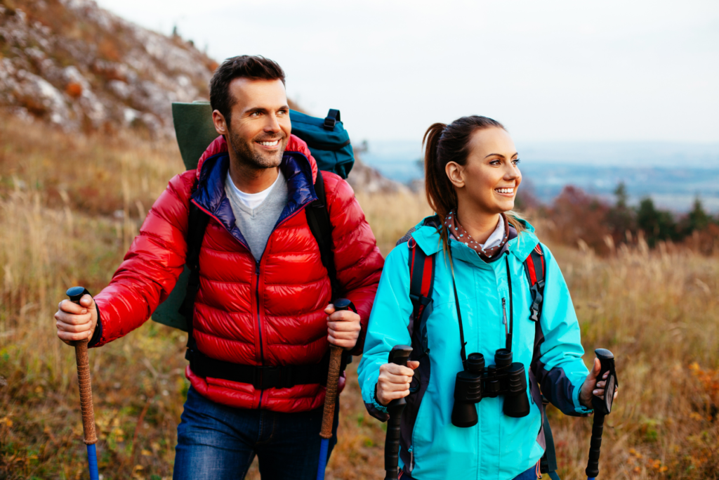 Image of man in red jacket and woman in blue jacket hiking through tall grass. Both are carrying hiking poles and looking off to the left. The woman has binoculars around her neck.