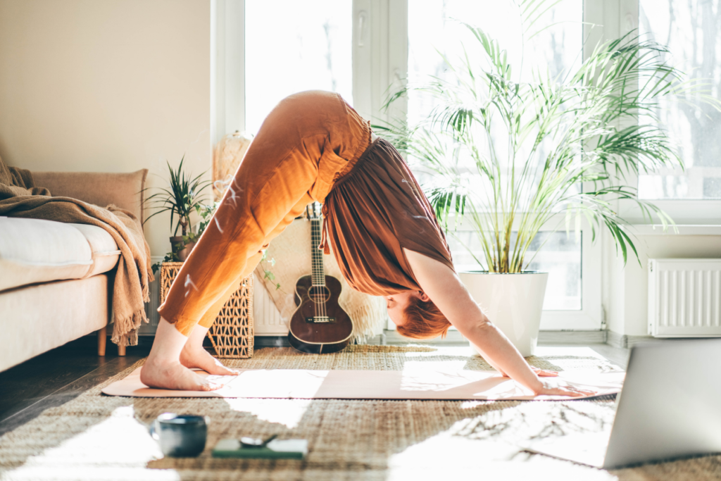 Image of active woman in a downward dog yoga pose in her living room. Woman is wearing burnt orange pants and a brown top. She is doing an at-home yoga class on her laptop. The living room is bright with the sun shining through the windows behind the woman.