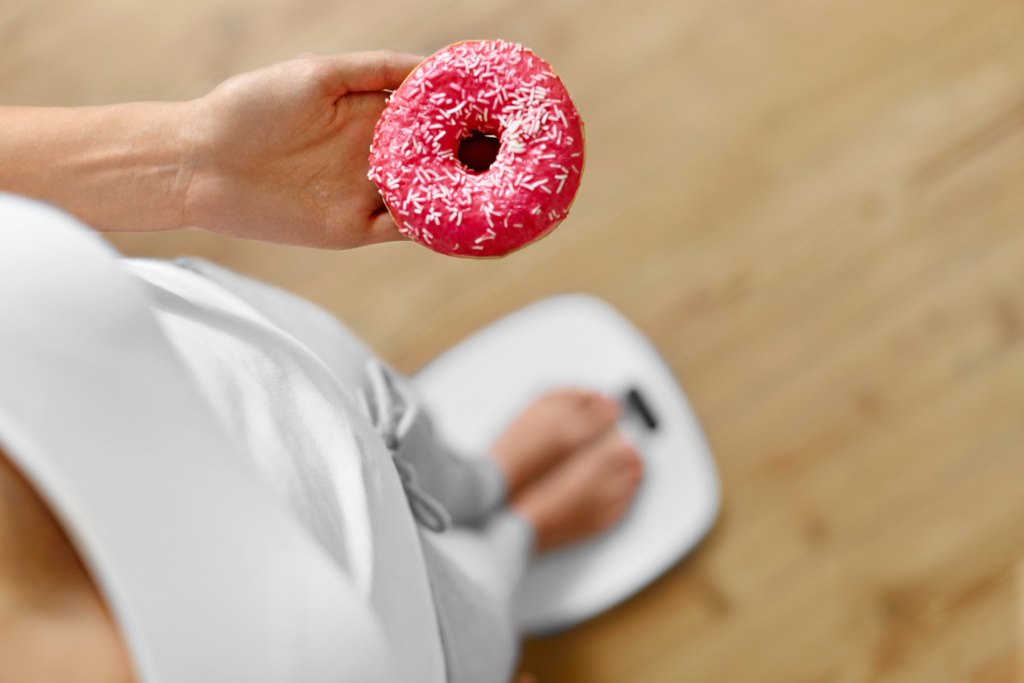 Overhead view of woman standing on a white weight scale and holding a doughnut