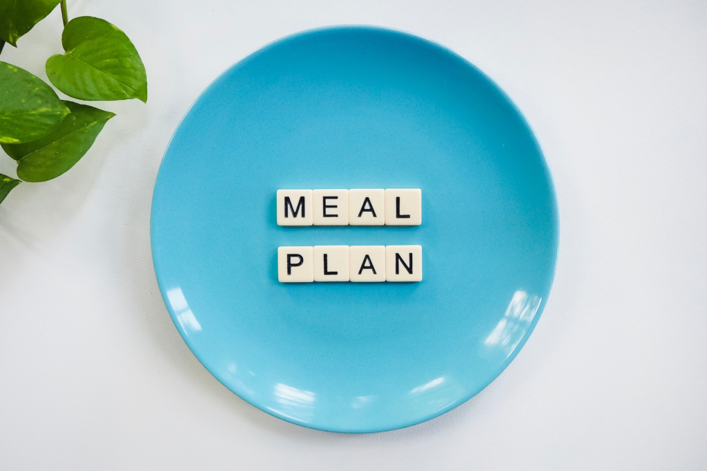 Blue plate on white background. The plate has scrabble letters on it that spell "meal plan". In the top left corner of the image are the green leaves of a plant.