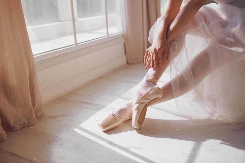 Image of a ballerina dancer's legs. the dancer is wearing pointe ballet shoes and has her ankles crossed and toes pointed. The dancer is sitting in-front of a bright window with curtians.