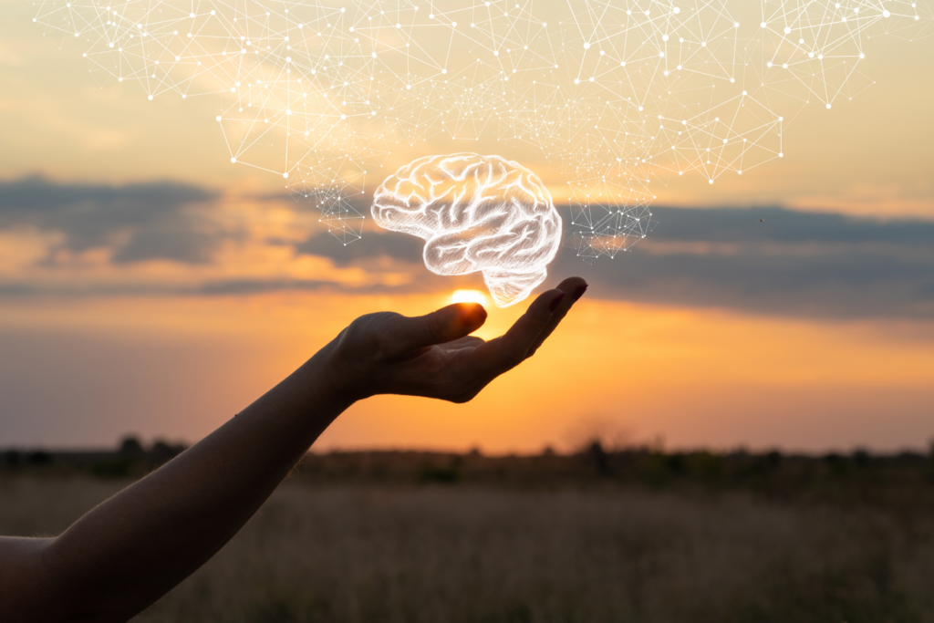 Image of hand held out with a hologram of a brain hovering above the palm. The image was taken outside with and orange sunset in the background