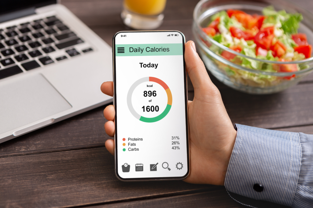 Image of hand holding phone with a calorie counting app opened reading 896 calories eaten out of 1600 calories for the day. There is a laptop, salad, and glass of orange juice in the background.