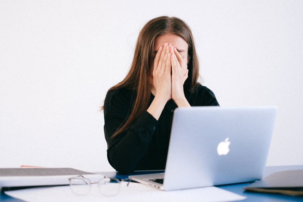 Image of a woman with brown hair wearing a black sweater covering her face with her hands. The woman is looking at a Macbook and appears to be stressed.