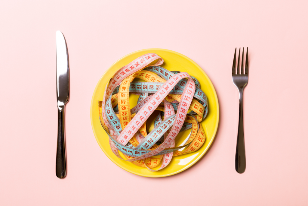 An image of a yellow plate covered in colourful measuring tapes with a fork and knife alongside it. The plate is on a pink background.