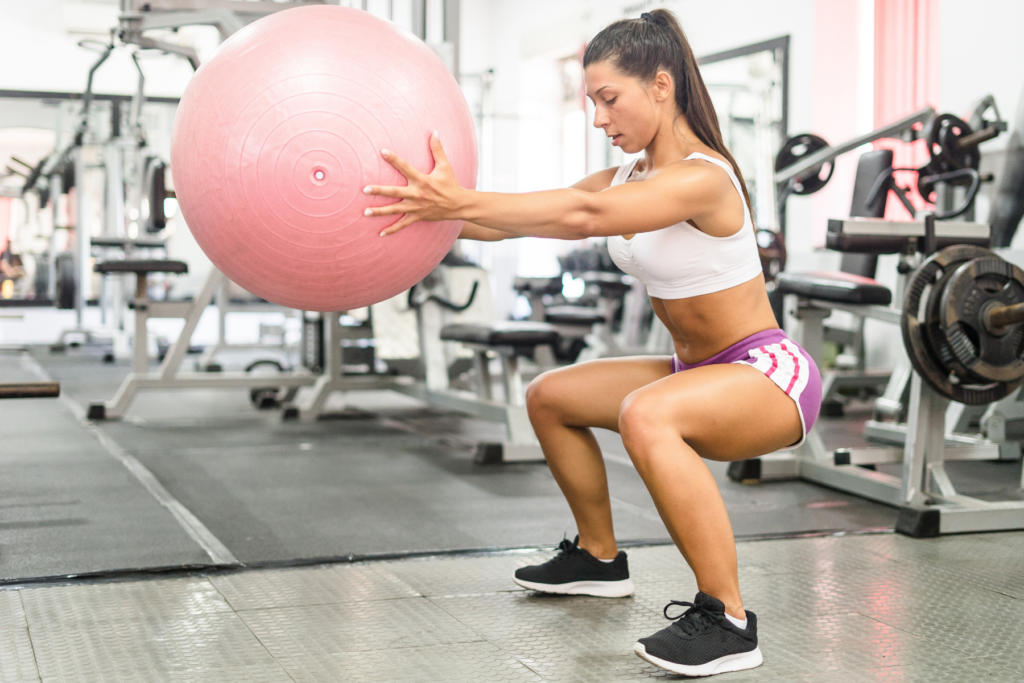 An image of a woman in a sports bra and athletic shorts at the gym. The woman is doing a squat with a pink exercise ball held out in front of her.
