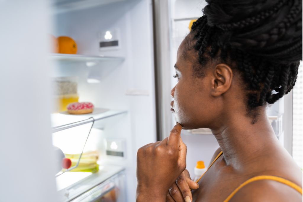 An image of a woman looking into an open fridge. The woman has her hand to her chin, pondering what she will choose to eat.
