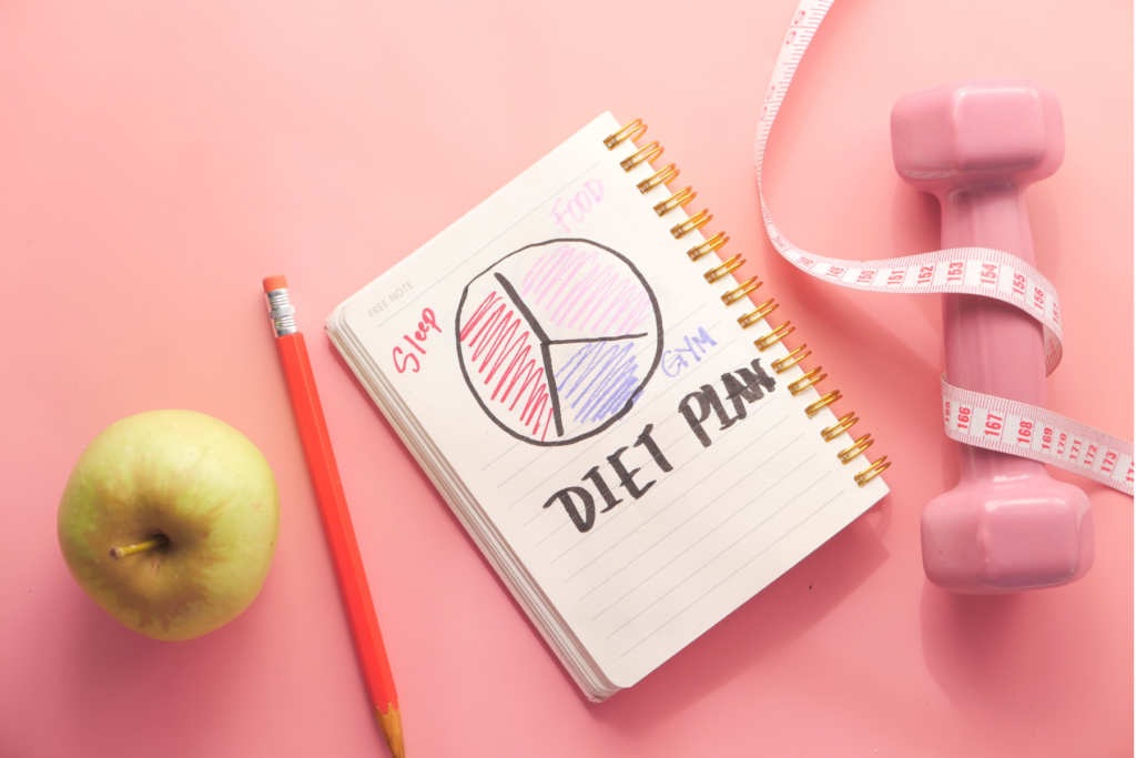 Overhead view of a notebook, green apple, pink dumbbell wrapped in a pink measuring tape, and a pencil on a pink surface. The notebook is folded open to a page that has a handdrawn pie grapgh and the words "diet plan" underneath.