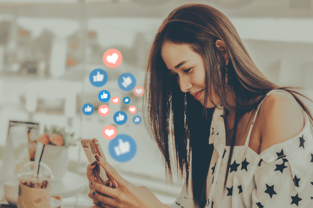 Woman looking down at an iphone that is in her hands. The woman is on social media. Blue bubbles with thumbs up icons and pink bubbles with heart icons are coming out of the phone.