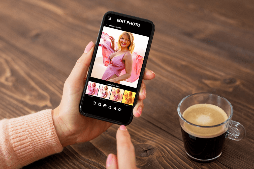 A woman's hands are holding an iphone above a wooden table on which sits a cup of coffee. The iphone is open to a photo editing app where an image of a woman in a pink dress is being edited with a filter.