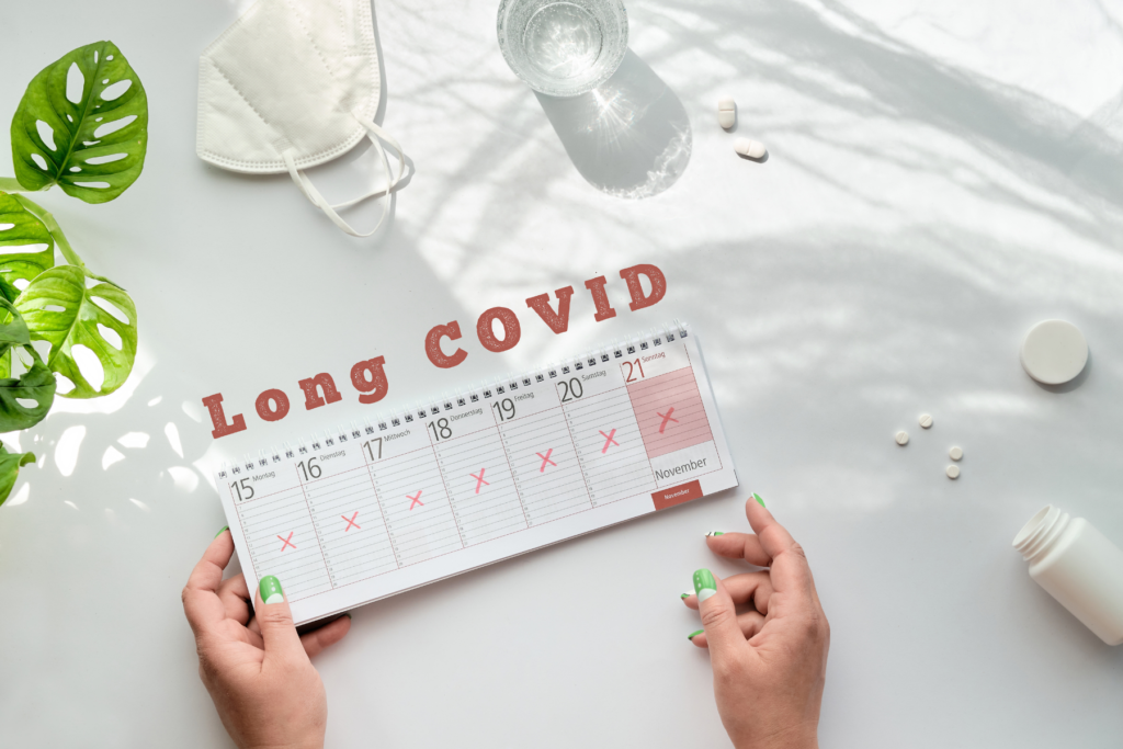 Calendar with red letter X through the days of the week. The words Long Covid in red above the calendar. The calendar is on a table with pills, a glass of water, and a medical mask.