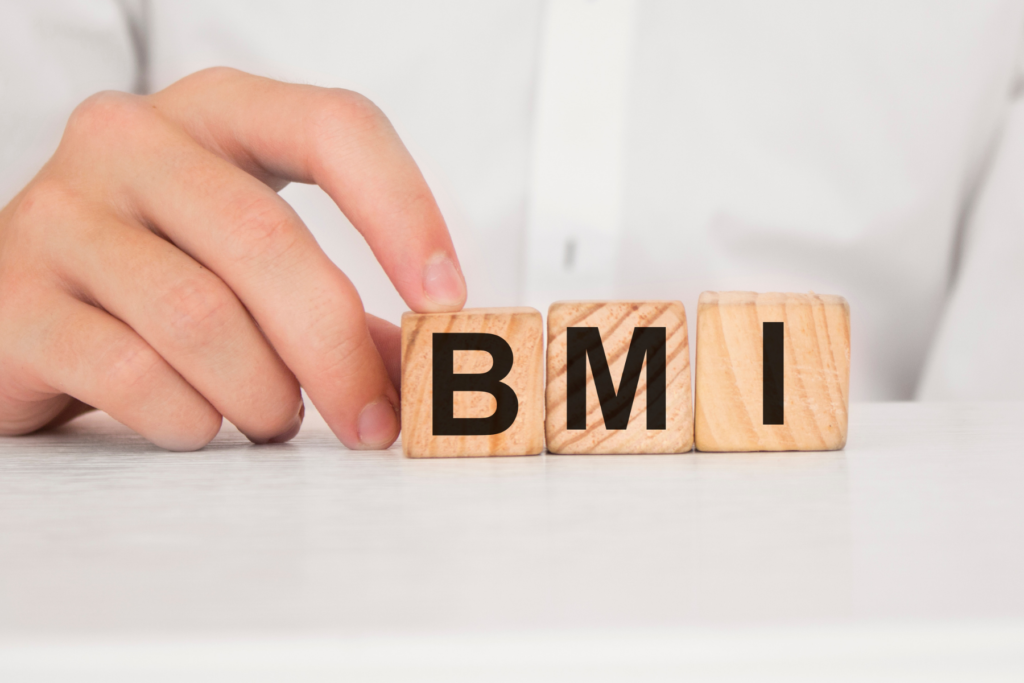 Three wooden letter blocks lined up side by side to spell BMI. The blocks sit on a white surface in front of a man's torso. The man wears a white button up and is touching the letter B block with one of his hands.