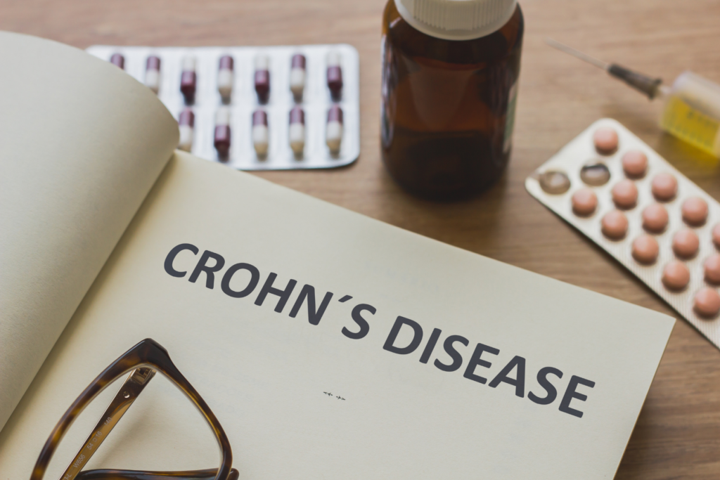 Picture of open notebook with the words Crohn's disease on a table surface. The book has a pair of reading glasses on it. On the table is also medication that is blister packed, a needle, and a medication bottle. 