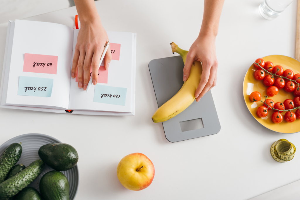 Overhead view of a countertop. A pair of hands is placing a banana on a food scale and recording calories in a notebook. The counter is covered in other foods including an apple, tomatoes, cucumbers, and avocados.