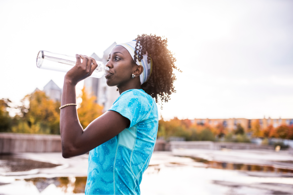 Black woman in blue activewear shirt and colourful headband standing outside in the fall. The woman has a clear water bottle to her mouth as she takes a drink for hydration while facing the left.