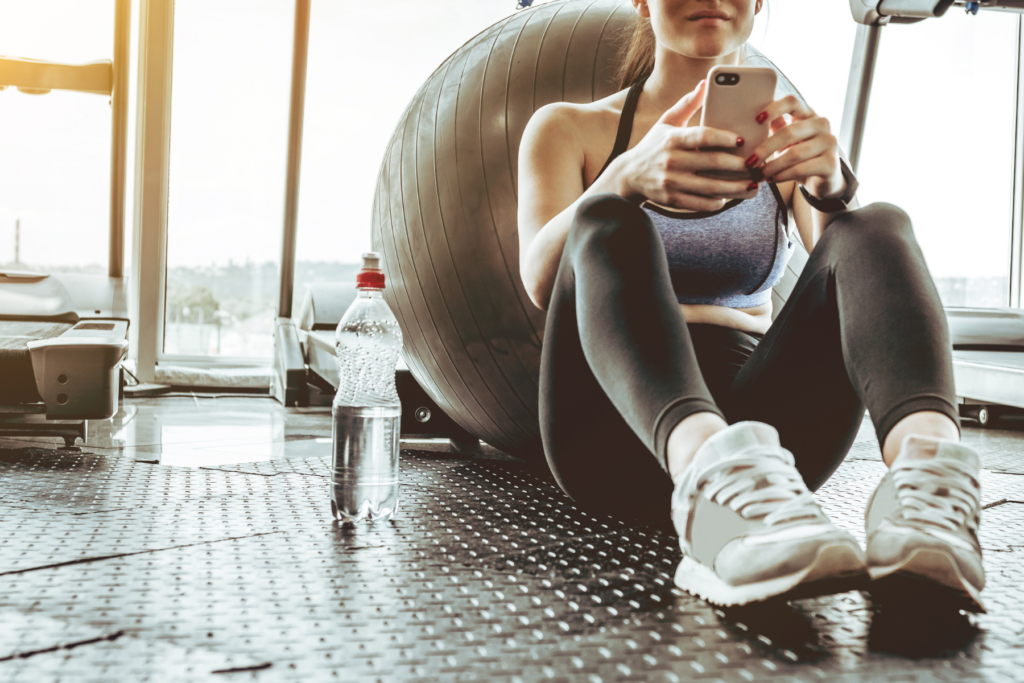 Woman sitting on a gym floor with her back against a grey workout ball. THe woman is on her phone. Beside her sits a clear, plastic water bottle for hydration.