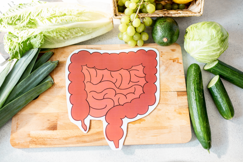 A wooden cutting board lays on top of a white kitchen countertop. Green vegetables and fruits surround the cutting board including lettuce, cucumber, grabes, and avocado. On top of the cutting board sits a piece of paper with a cut out drawing of the intestines and colon.