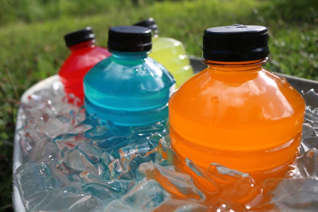 Four bottles of sports drinks or 
 hydration electrolyte drinks inside a tub of ice outside. The drinks are coloured orange, blue, red, and yellow.
