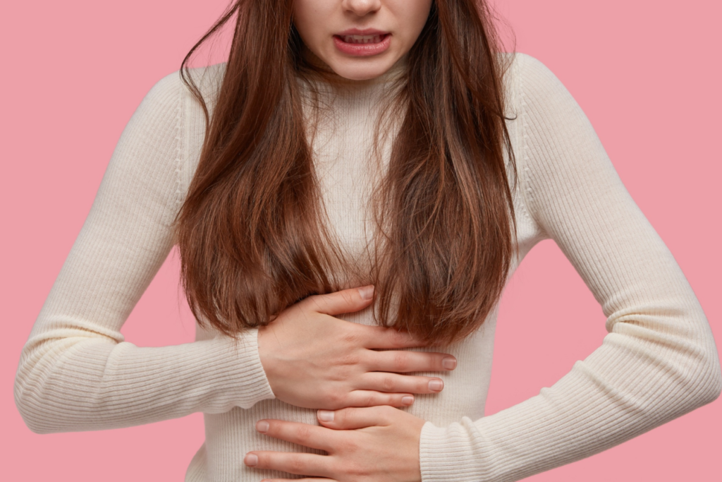 Woman with in a while sweater and with brown hair holding her hands to her stomach. The woman's teeth are clenched like her stomach is in pain. The woman stands in front of a pink background.