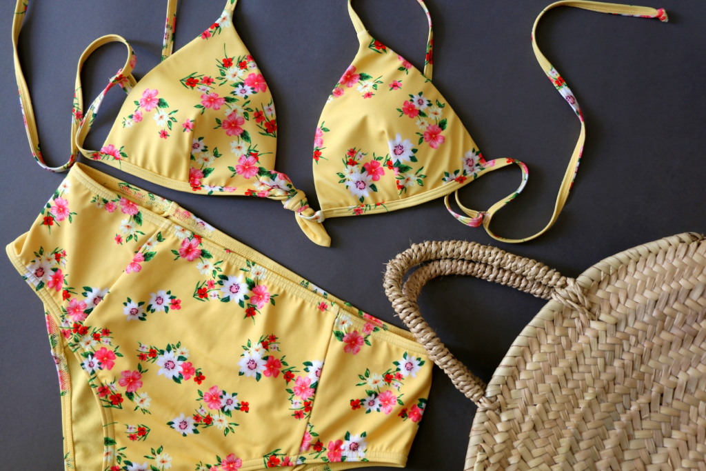 Overhead view of of a yellow beach bikini with pink and white flowers lain out on a black background. In the right bottom corner of photo is a wicker purse