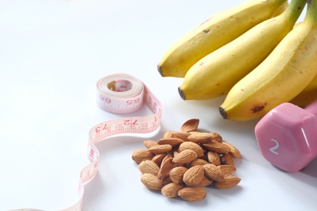 Image with a bunch of bananas  a pink 2lb dumbbell, a pile of almonds, and a flexible measuring tape.