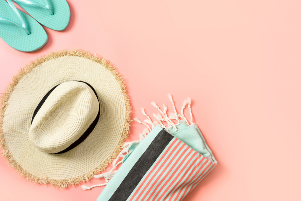 Image of straw sun hat, turquoise summer flip flops, and a turquoise beach towel with pink stripes on a blush pink background.