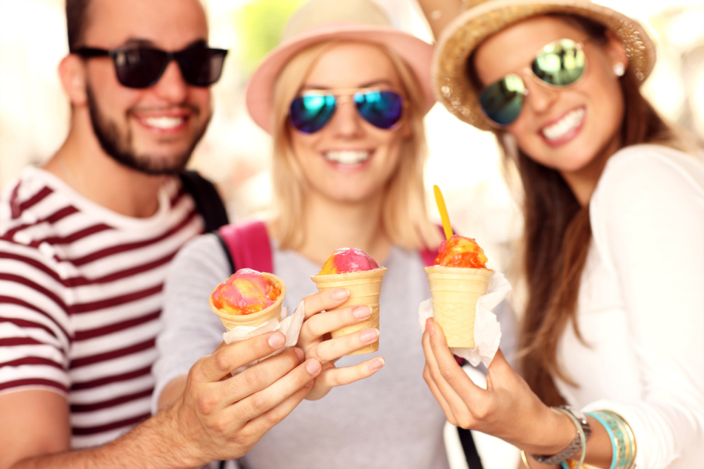 Image of three friends standing together, one man on the left and two women on the right. The friends are wearing sunglasses and their faces and bodies are out of focus. In the foreground the friends each hold up an ice cream, cone of pink and orange sherbert. 