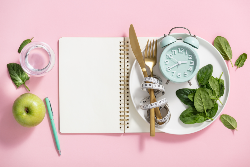 Image of an open notebook with blank white pages on a light pink surface. On the right side side page of the notebook sits a white plate that is topped with spinach leaves, a mint green alarm clock and a gold knife and fork that are tied together with a white measuring tape. To the left of the notebook sits a glass of water, a green apple, a mint green pen and more spinach leaves. The image represents dieting weight loss. 