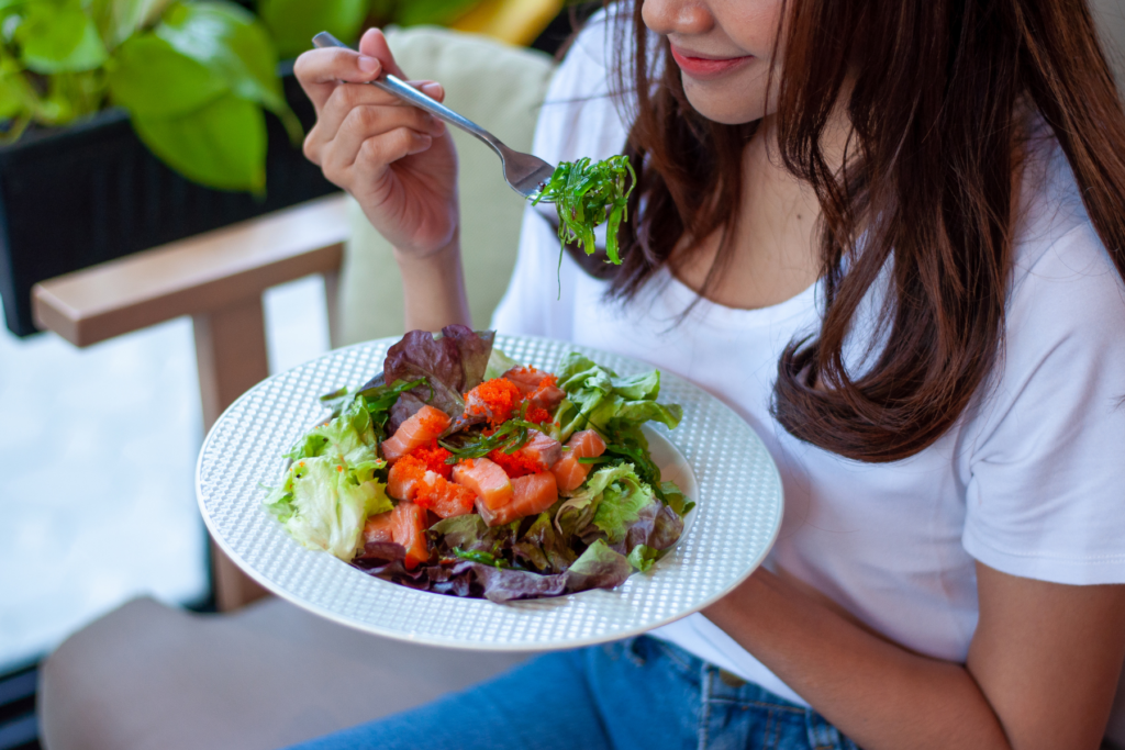 Image of a woman in blue jeans and a white shirt sitting in a cushioned chair. The top of the woman's head is out of the frame. The woman is holding a bowl of salad that has lettuce and tomatoes. The woman is smiling as she holds a forkful of salad in her right hand up to her mouth. 
