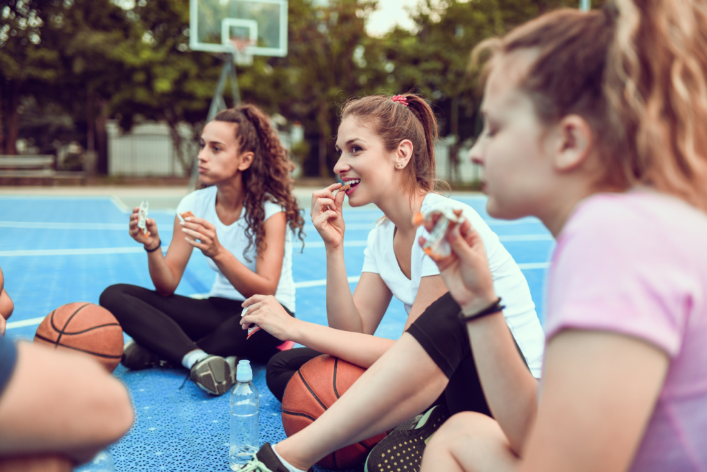 Image of three young female basketball players sitting on the ground of an outdoor basketball court. The girls are sitting cross legged and are eating a snack of granola bars.