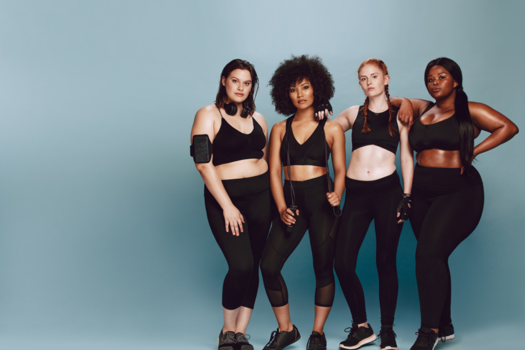 Image of four women athletes in black leggings and sports bras. The women are of different ethnicities and of different body types. The women stand in front of a blue background in a row with their arms resting on each other's shoulders. 