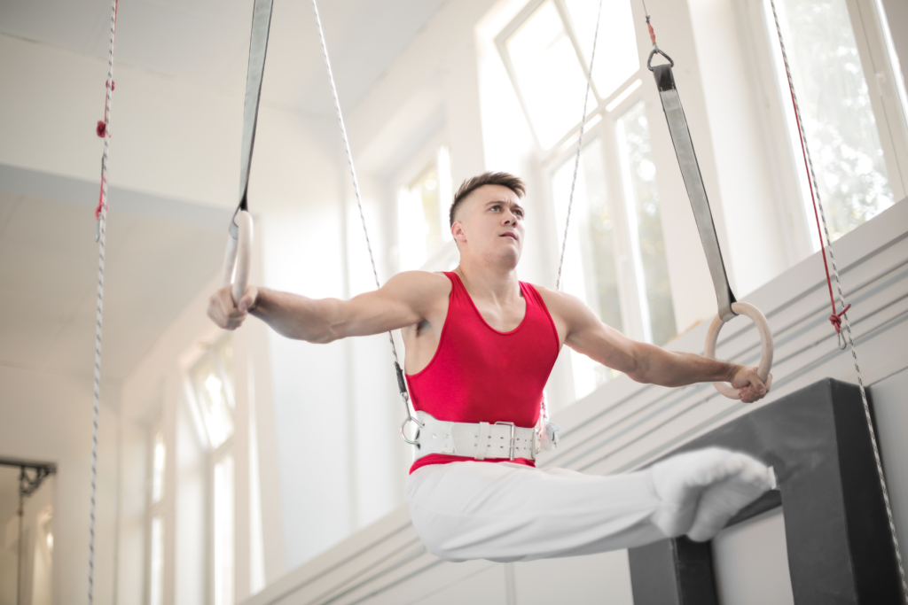 Image of a male gymnast in a red tank top and white pants. The gymnast holds himself up on the rings by extending his arms by his sides. The gymnast holds his legs straight out infront of himself in a pike position.
