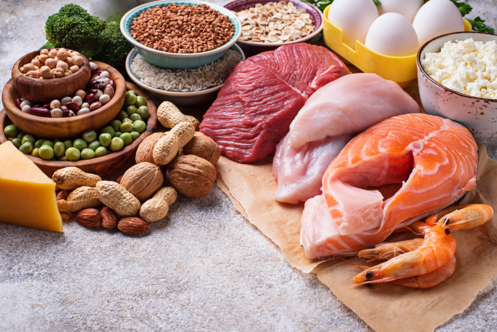 Image of various food sources of protein on a kitchen counter. Included in the image is meats like chicken and steak, seafood like salmon and shrimp, legumes like peanuts and beans, grains like rice and oats, as well as eggs and cheese.