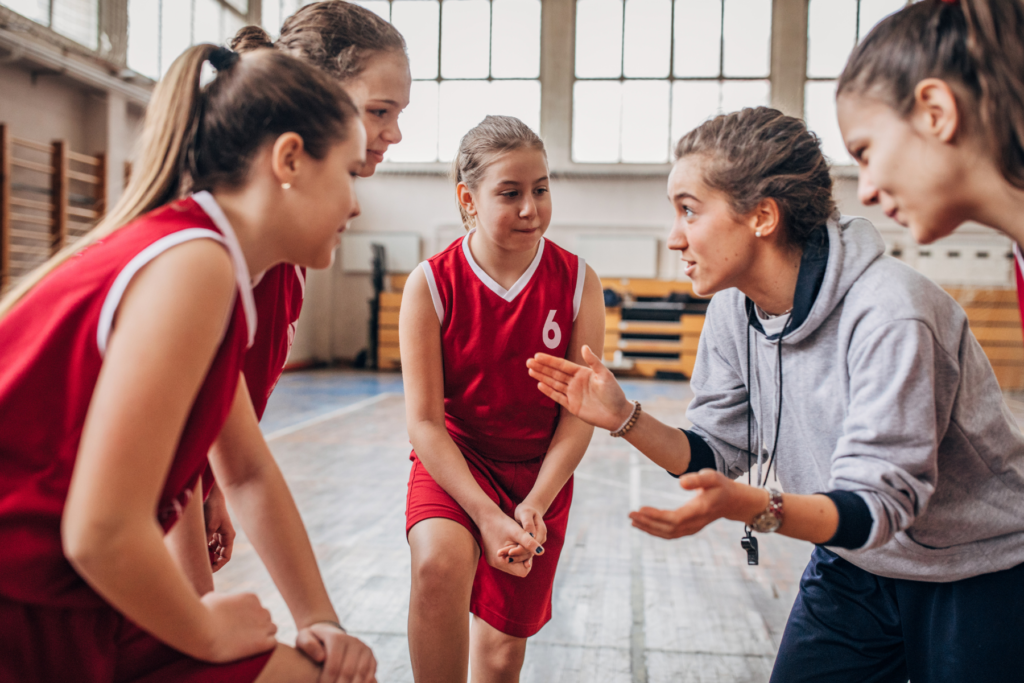 Female sports coach in grey sweatshirt talking intently with her young, female athletes. The athletes are wearing red team uniforms and are huddled in a circle with the coach.