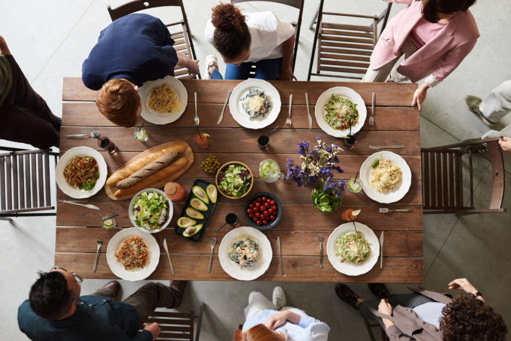 Birds Eye view of eight people gathering and starting to take seats around a large, wooden dining table. THe table is set with plates of pasta and cutlery. In the centre of the table is a vase of flowers, loaves of crusty bread, salads, and avocados that are cut in half.