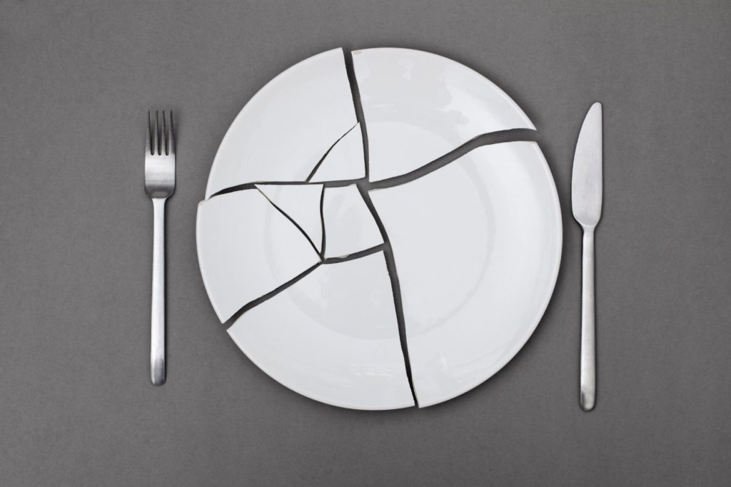 Image of a while plate broken into pieces to represent eating disorders. The pieces of the plate are placed on a grey background as if someone tried to put them back together again. On the left side of the plate is a silver fork and on the right, a silver knife.