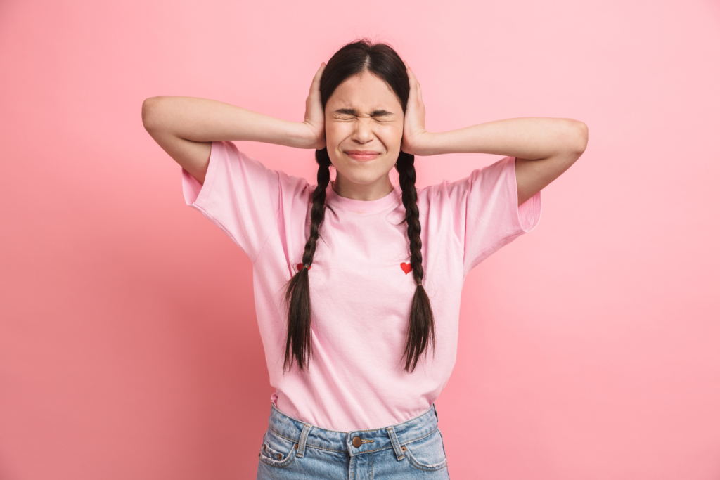 A young woman with a pink t-shirt and blue jeans stands in front of a pink wall. The girl two, long brown braids. She holds her hands up to her ears and squints her eyes closed as if she is trying to block out sound or ignore somebody.