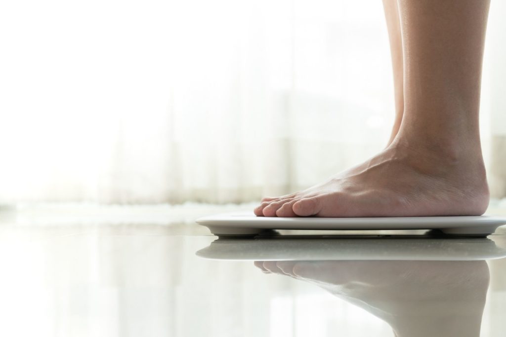 Image of a pair of bare feet pointing towards the right and standing on a white bathroom scale to measure weight.
